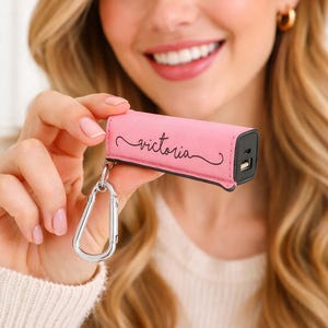 Pink personalized power bank keychain charger with engraved name, compact portable phone charger clipped to keys and charging a smartphone, travel friendly tech accessory gift.