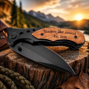 May include: A black folding knife with a wooden handle engraved with "Sharpest Dad Around Est. 2026" rests on a wooden surface. The blade is open, revealing a sharp, black edge. The background features a scenic view of mountains and a sunset.