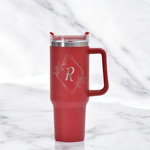 May include: A red insulated tumbler with a handle and a straw. The tumbler features a silver floral design with the letter "R" inside a diamond shape. The lid is silver and has a red slider. The tumbler is designed to keep drinks cold or hot.