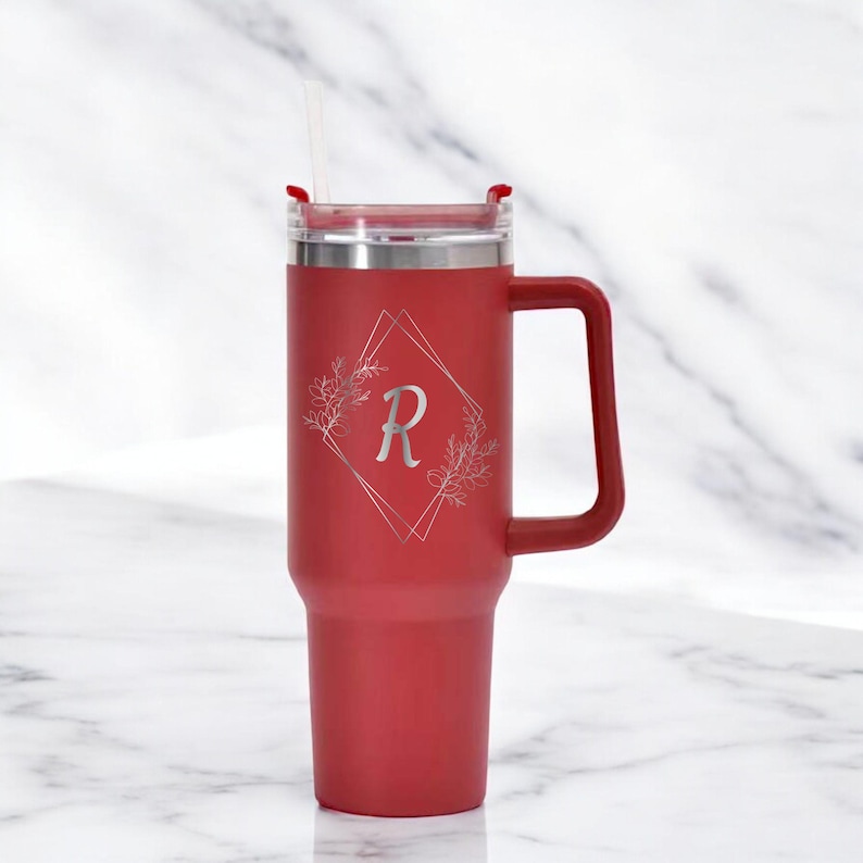 May include: A red insulated tumbler with a handle and a straw. The tumbler features a silver floral design with the letter "R" inside a diamond shape. The lid is silver and has a red slider. The tumbler is designed to keep drinks cold or hot.