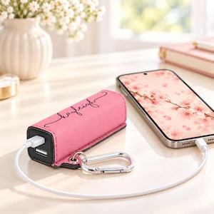 May include: A rectangular pink portable power bank with a black trim and a silver metal carabiner attached to the side. The power bank features the name Kayleigh written in black cursive script on the top surface and is connected to a smartphone.