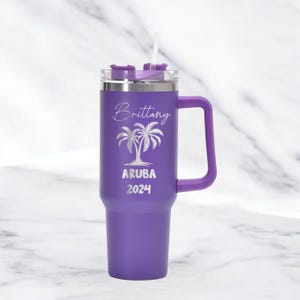 May include: A purple travel mug with a handle and a straw. The mug has the name "Brittany" and the words "ARUBA 2024" with a palm tree design. The mug is designed to hold a beverage.
