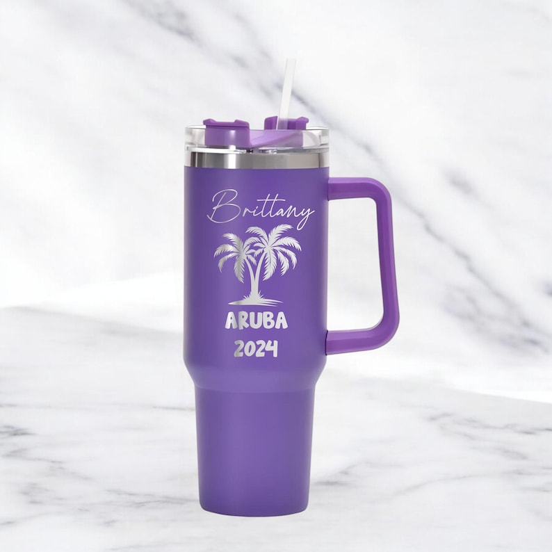May include: A purple travel mug with a handle and a straw. The mug has the name "Brittany" and the words "ARUBA 2024" with a palm tree design. The mug is designed to hold a beverage.