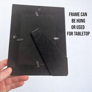 May include: Black picture frame with a stand on the back. The frame can be hung or used for tabletop display. "FRAME CAN BE HUNG OR USED FOR TABLETOP" is printed on the back of the frame.