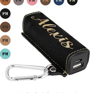 May include: Black leather phone charger case with a gold inscription that reads "Alexis". The case has a yellow stitch detail and a silver carabiner clip.