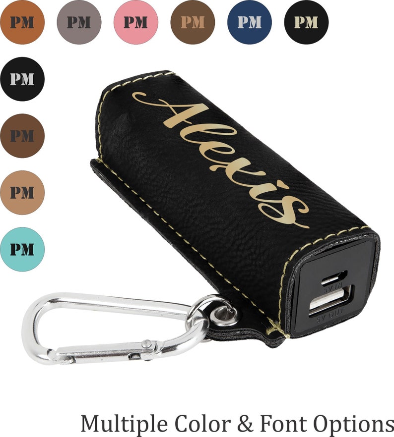 May include: Black leather phone charger case with a gold inscription that reads "Alexis". The case has a yellow stitch detail and a silver carabiner clip.