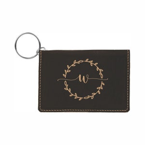May include: A brown leather key chain with a key ring and a small pocket for cards. The pocket has a gold-colored monogram of the letter "W" inside a wreath of leaves.