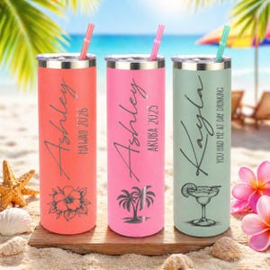May include: Three colorful stainless steel tumblers with straws on a sandy beach. One is coral with "Ashley Hawaii 2026", another is pink with "Ashley Aruba 2025", and the last is mint green with "Kayla You had me at day drinking".