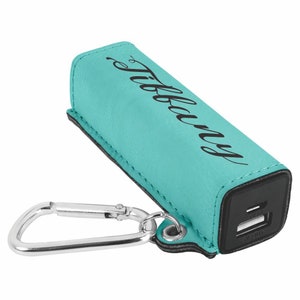 May include: A turquoise leather power bank with a black carabiner clip and the name "Tiffany" engraved on the front. The power bank has a black USB port on the side.