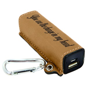 May include: A brown leather-covered portable phone charger with a carabiner clip. The charger has a black USB port and the text "You are the charge in my heart" is engraved on the front.