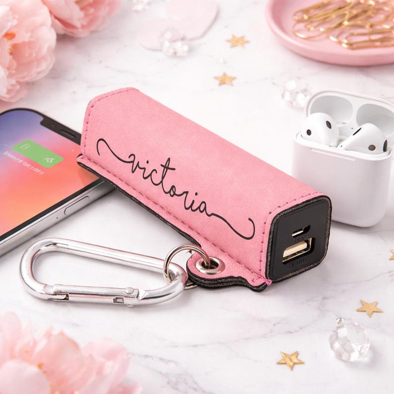 Custom engraved portable phone charger keychain with pink leatherette cover and name engraving, small power bank charging a smartphone with keys and earbuds nearby, personalized tech gift idea for her.
