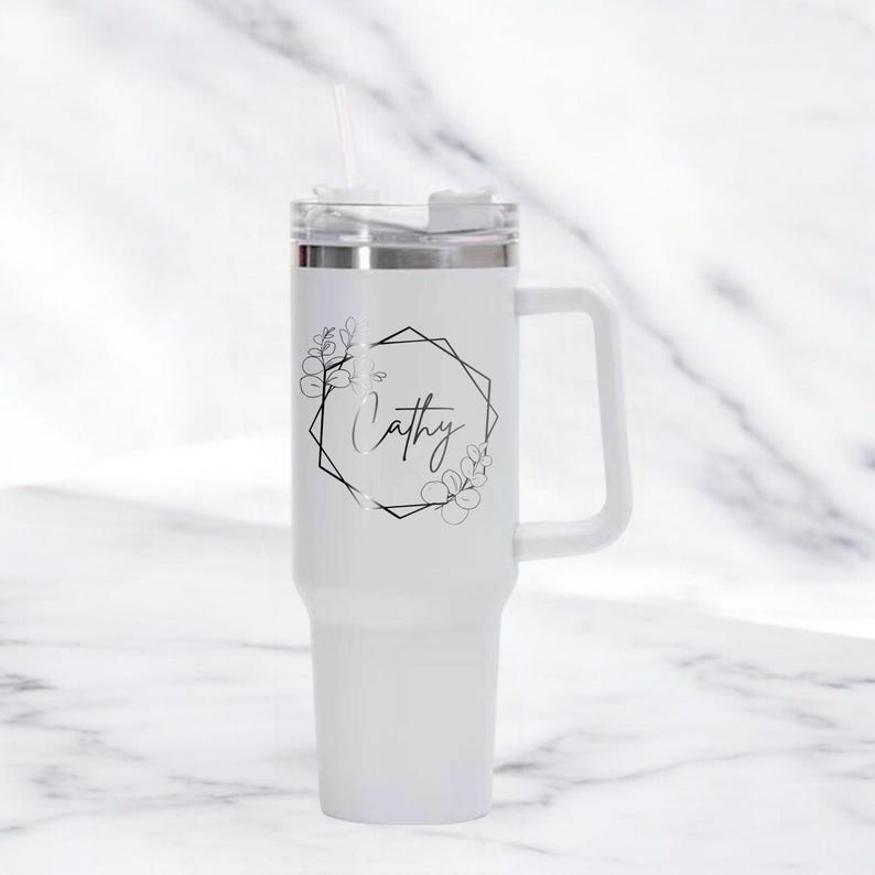 May include: White insulated tumbler with a handle and a straw. The tumbler features a black floral design around a geometric frame with the name "Cathy" in script. The lid is silver and white.