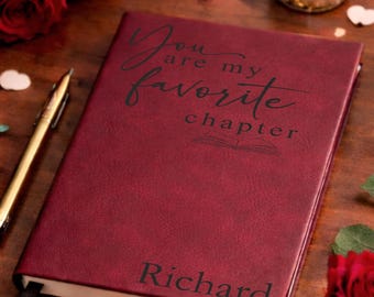 May include: A burgundy journal with the inscription "You are my favorite chapter" and the name "Richard" is displayed on a wooden surface. Red roses, a lit candle, and decorative hearts surround the journal, creating a romantic ambiance.