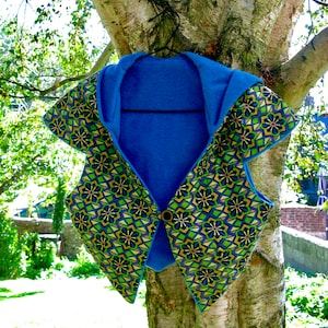 Peut inclure: Un gilet à capuche bleu avec un motif géométrique dans des tons de vert, or et bleu. Le gilet est suspendu à une branche d'arbre.