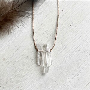 May include: A clear quartz crystal pendant necklace. The pendant features several clear quartz crystal points, suspended from a thin, brown cord. The necklace is displayed on a white, textured surface, with a feather in the upper left corner.