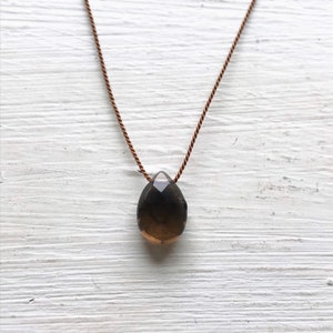 Puede incluir: Un collar con colgante de cuarzo ahumado en forma de lágrima. La gema facetada, de color marrón oscuro, cuelga de un delicado cordón marrón. El collar se exhibe sobre un fondo de madera blanca.