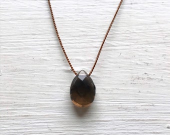 May include: A teardrop-shaped smoky quartz pendant necklace. The faceted, dark brown gemstone hangs from a delicate, brown cord. The necklace is displayed against a white, wooden background.