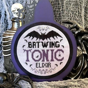 May include: A purple, bottle-shaped decoration with a circular design featuring a bat and the words "Batwing Tonic Elixir." The design is in black and purple, with a spider and other decorative elements. A skull and spider are in the background.