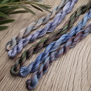 May include: Six skeins of embroidery thread in shades of blue, green, and brown are displayed. The threads are twisted and arranged diagonally. The colours vary from pale blue to dark indigo, with touches of green and brown. The threads are on a wooden surface.