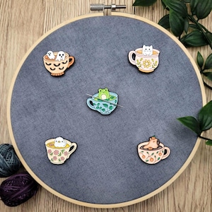 May include: Five enamel pins featuring creatures in teacups. The pins show a ghost, a cat, a frog, and a bear. The teacups are in orange, yellow, and blue, set on grey fabric stretched over a wooden embroidery hoop. The pins are decorative.