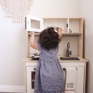 May include: A child in a blue dress plays with a wooden toy kitchen. The kitchen has a white cabinet, a sink, a tap, and a microwave. A towel with the text "AUTUMN'S KITCHEN" hangs on the front.