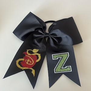 May include: Black cheerleading bow with a red and gold glitter snake and apple design on one tail and a green glitter letter "Z" on the other. The bow is made of a wide, ribbed fabric.