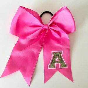 Puede incluir: Un lazo de animadora rosa brillante con un diseño llamativo. El lazo presenta una gran letra "A" en purpurina verde con un borde blanco. Está sujeto a una goma elástica negra para el pelo.