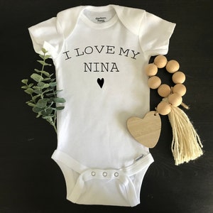 May include: White baby onesie with black text that reads "I love my Nina" with a small black heart below the text. The onesie is lying on a black surface with a wooden bead garland and a wooden heart.