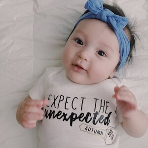 May include: A baby wearing a white onesie with the text "Expect the Unexpected" and a key with the text "Autumn 8/19". The baby is wearing a blue headband.