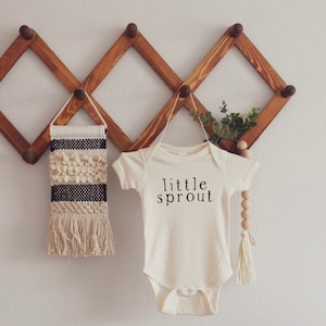 Op de afbeelding: Een witte baby romper met de tekst "little sprout" in zwart gedrukt. De romper hangt aan een houten rek met een klein geweven wandkleed en een houten kralenketting.