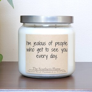 May include: A white soy candle in a glass jar with a silver lid. The candle has a brown label with the text "I'm jealous of people who get to see you every day." and "The Southern Flame Signature Blend | Premium 16 oz. Soy Candle".