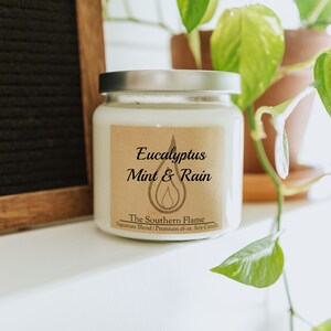 May include: A white soy candle in a clear glass jar with a silver lid. The label reads "Eucalyptus Mint & Rain" and "The Southern Flame." The candle is 16 oz.