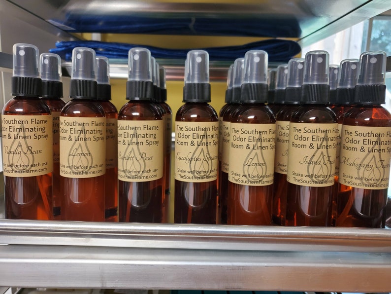 May include: A collection of brown glass spray bottles filled with liquid. Each bottle has a black spray nozzle and a label with text. The labels read "Southern Flame Odor Eliminating Room & Linen Spray" with various scents like Vanilla Bean and Lemon.