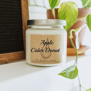May include: A white soy candle in a glass jar with a silver lid. The label reads "Apple Cider Donut" with a droplet design. The candle is 16 oz. and is part of The Southern Flame Signature Blend.
