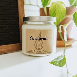 May include: A white soy candle in a glass jar with a silver lid. The label reads "Gardenia" above "The Southern Flame" and "Premium 16 oz. Soy Candle". The candle is on a white surface with green plants in the background.