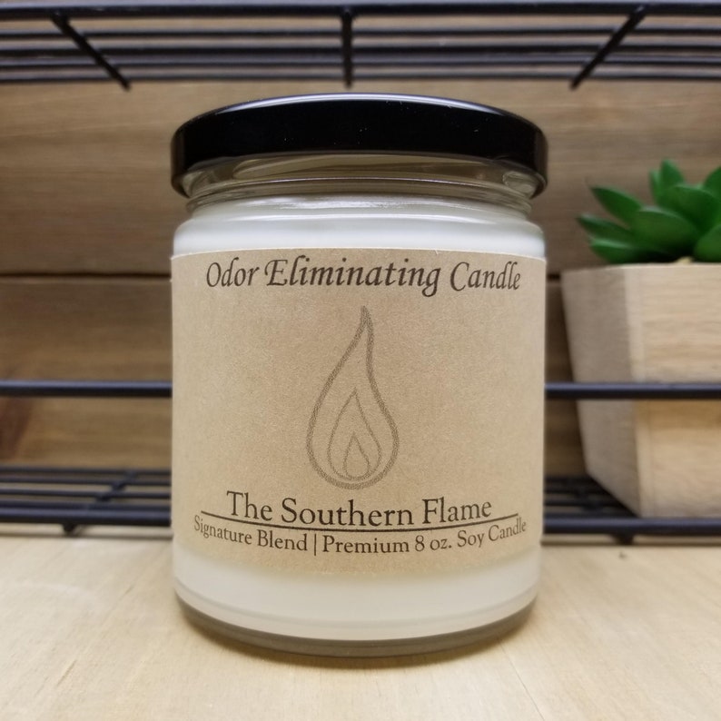 May include: A clear glass jar candle with a black lid. The label reads "Odor Eliminating Candle" and "The Southern Flame." The candle is a soy blend and is 8 oz. The label features a flame graphic.
