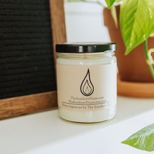 May include: A white candle in a clear glass jar with a black lid. The jar has a brown paper label with the text "TheSouthernFlame.com TheSouthernFlame.etsy.com Home fragrances by The Southern Flame".