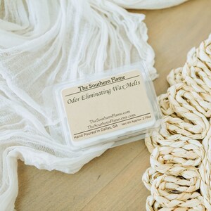 May include: A clear plastic container of wax melts with a beige label that reads "The Southern Flame Odor Eliminating Wax Melts." The container is on a white cloth and a woven straw mat. The net weight is approximately 2.75 ounces.
