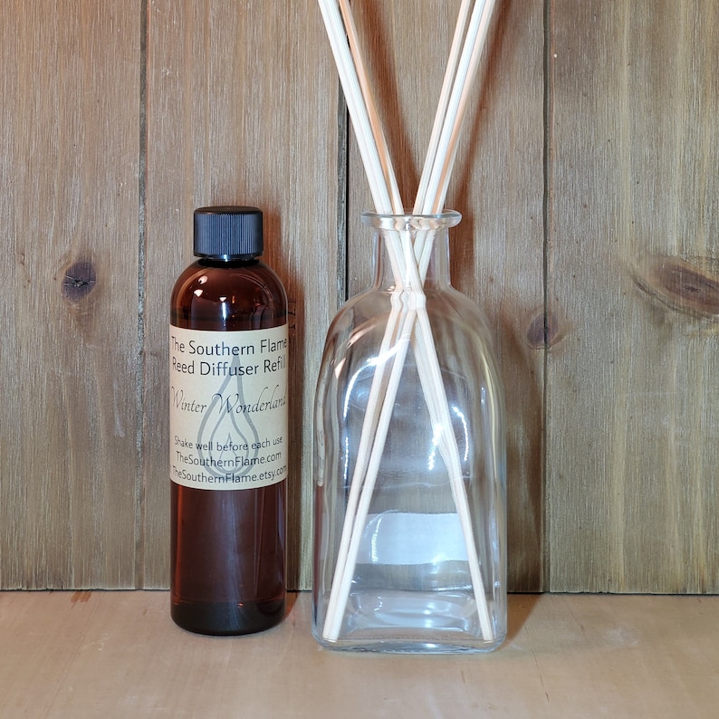 May include: A clear glass reed diffuser bottle with wooden reeds and a brown bottle of reed diffuser refill. The refill bottle has a black cap and a label that reads "The Southern Flame Reed Diffuser Refill Winter Wonderland."