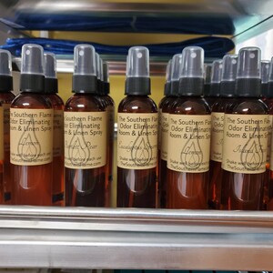 May include: A shelf display of brown glass spray bottles with various scents, including Vanilla Bean, Lemon,  Ripe Pear, Eucalyptus, Island Fresh, and Mahogany. Each bottle has a white label with the text "The Southern Flame Odor Eliminating Room & Linen Spray" and the scent name.