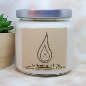 K&ouml;nnte beinhalten: Eine wei&szlig;e Sojakerze in einem transparenten Glas mit silbernem Deckel. Das Etikett zeigt eine Flammen-Grafik und den Text "The Southern Flame Signature Blend | Premium 454g Soy Candle". Links ist eine kleine Sukkulente in einem Holztopf zu sehen.
