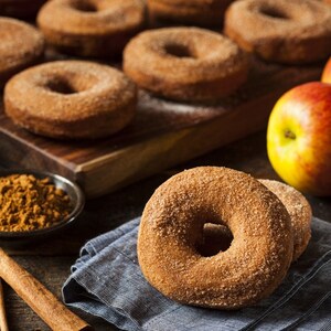May include: A close-up shot of several cinnamon sugar-covered donuts. The donuts are arranged on a wooden surface with a bowl of cinnamon and a red apple. A blue cloth and cinnamon sticks are also visible.
