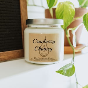 May include: A white soy candle in a glass jar with a silver lid. The label reads "Cranberry Chutney" with a flame graphic and "The Southern Flame" branding. The candle is 16 oz.