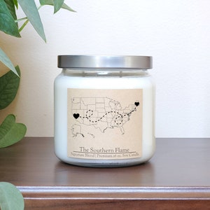 May include: A white soy candle with a brown label featuring a map of the United States with a dotted line connecting two hearts. The label reads "The Southern Flame Signature Blend | Premium 16 oz. Soy Candle".
