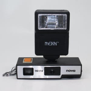 May include: A black and silver Nova 110 FX camera with a black Meikai flash unit attached. The camera has an orange button and a silver chain. The flash unit has the brand name "MEIKAI" printed on it.