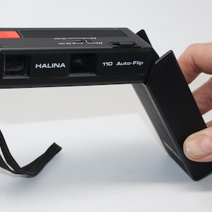 May include: Black Halina 110 Auto-Flip camera with a red button and a black wrist strap. The camera is rectangular with the brand name "HALINA" printed on the front.