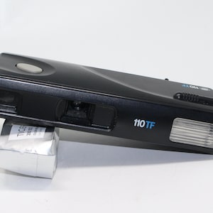 May include: A black 110TF film camera with a built-in flash. The camera has a silver button on the top and a small viewfinder on the front.