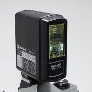 Könnte beinhalten: Ein schwarzer National PE-145 Kamera-Blitz mit gelber Linse. Der Blitz hat eine rechteckige Form mit einem Bedienfeld. Der Text "National PE-145" ist sichtbar.