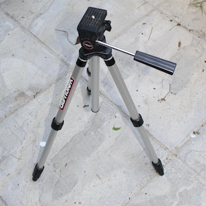 May include: A silver and black Vanguard tripod with three legs. The tripod has a black head with a handle and a mounting plate. The Vanguard logo is visible on one leg. The tripod is designed for photography and videography.