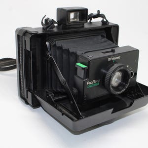 May include: A black Polaroid ProPack camera featuring a folding bellows design, a front-mounted lens with a circular focus ring, a viewfinder on top, and a green accent button. The camera body displays the text Polaroid and ProPack camera.